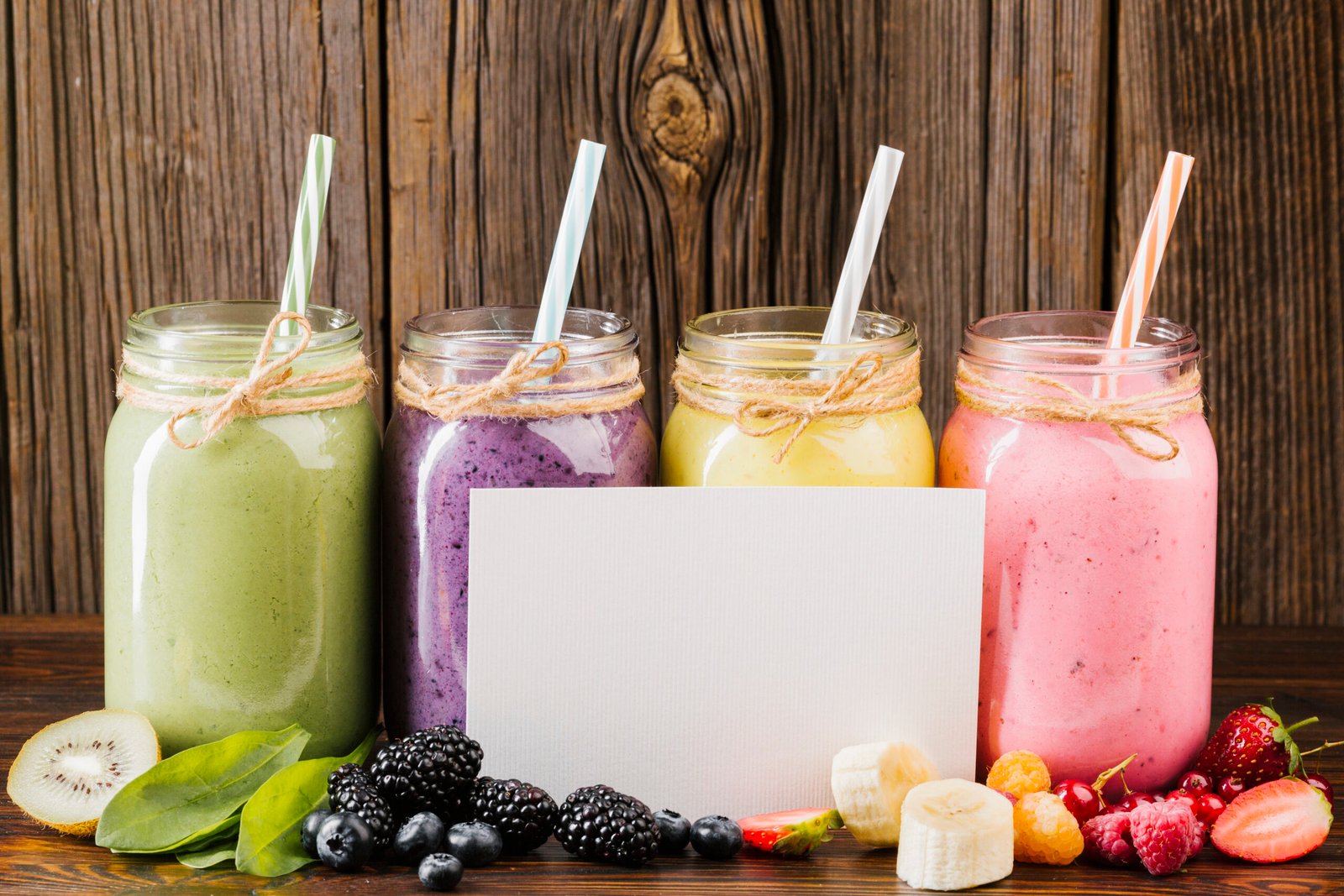 Cuatro frascos de vidrio con licuados de colores (verde, morado, amarillo y rosa) decorados con lazo de yute, junto a una tarjeta blanca vacía y frutas frescas sobre madera.