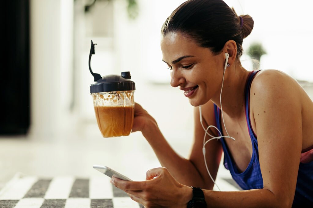 smiling woman drinking fruit smoothie while using mobile phone at home.