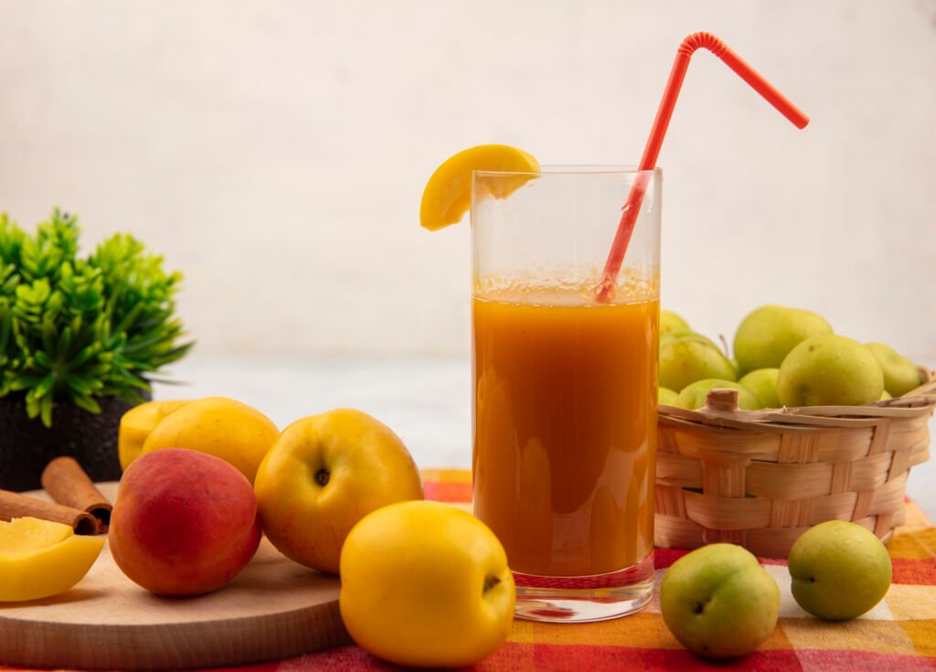 side view of sweet yellow peaches on a wooden kitchen board with pinky orange peach with peach juice with green cherry plums on a bucket on a checked tablecloth on a white background