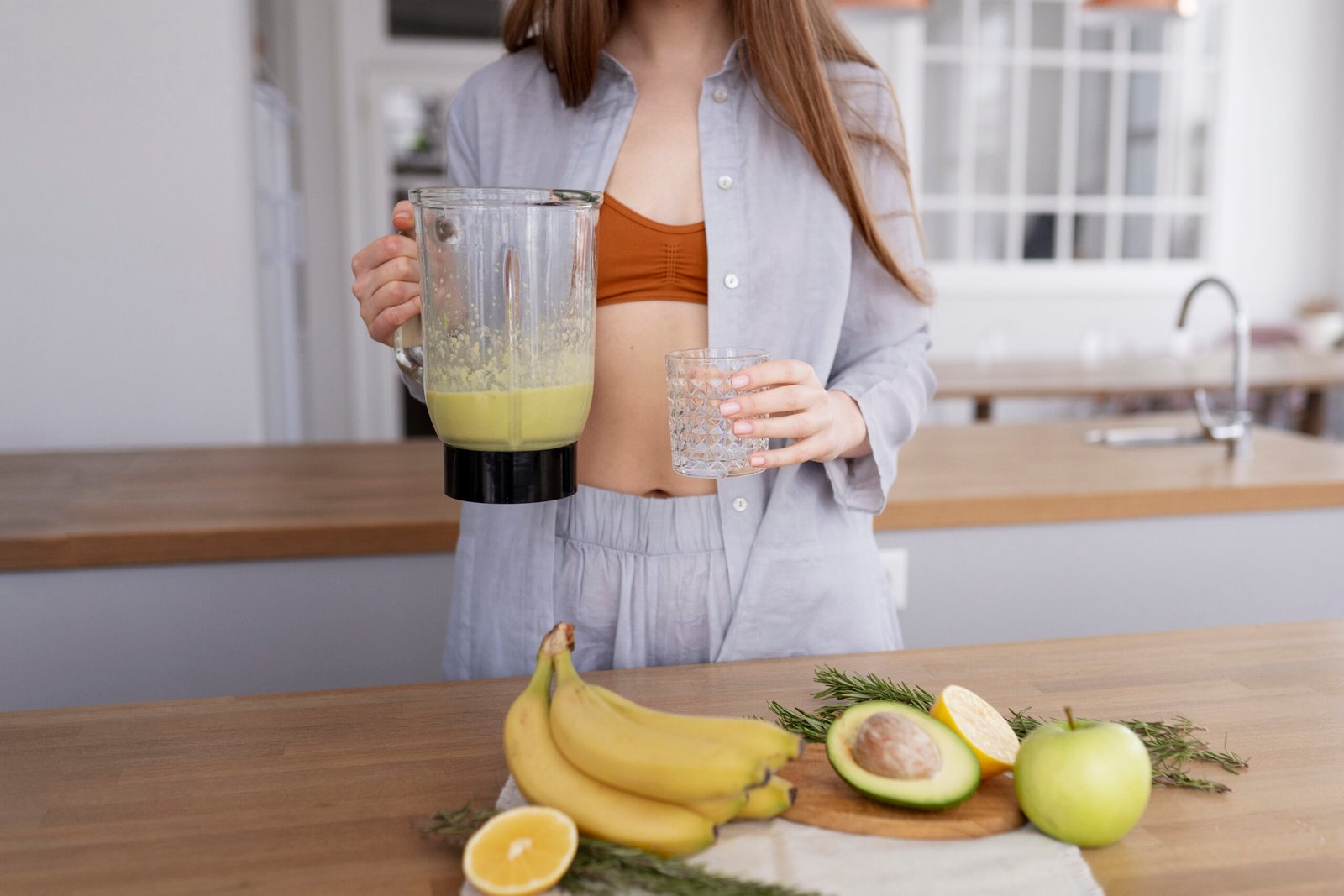 Mujer en la cocina sosteniendo una jarra de licuadora con batido verde y un vaso vacío, con plátanos, aguacate y manzana verde sobre la barra.