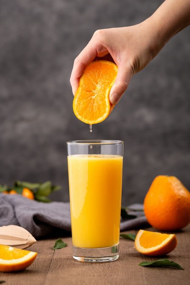 Una mano apretando media naranja sobre un vaso lleno de jugo fresco, capturando una gota cayendo, creando una bebida saludable sobre una mesa de madera con naranjas enteras y cortadas al fondo.