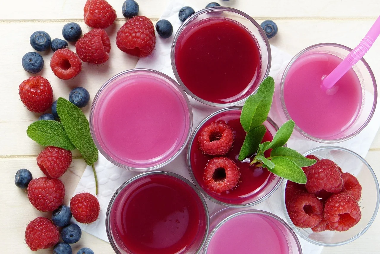 Vista aérea de varios vasos con jugos en tonalidades rosa y magenta, rodeados de frambuesas frescas, arándanos y hojas de salvia sobre una superficie de madera clara.