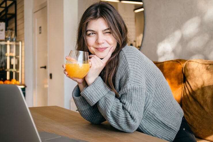 Una mujer joven con suéter gris sonríe y guiña un ojo mientras sostiene un vaso de jugo de naranja frente a una laptop en una cafetería iluminada por el sol.