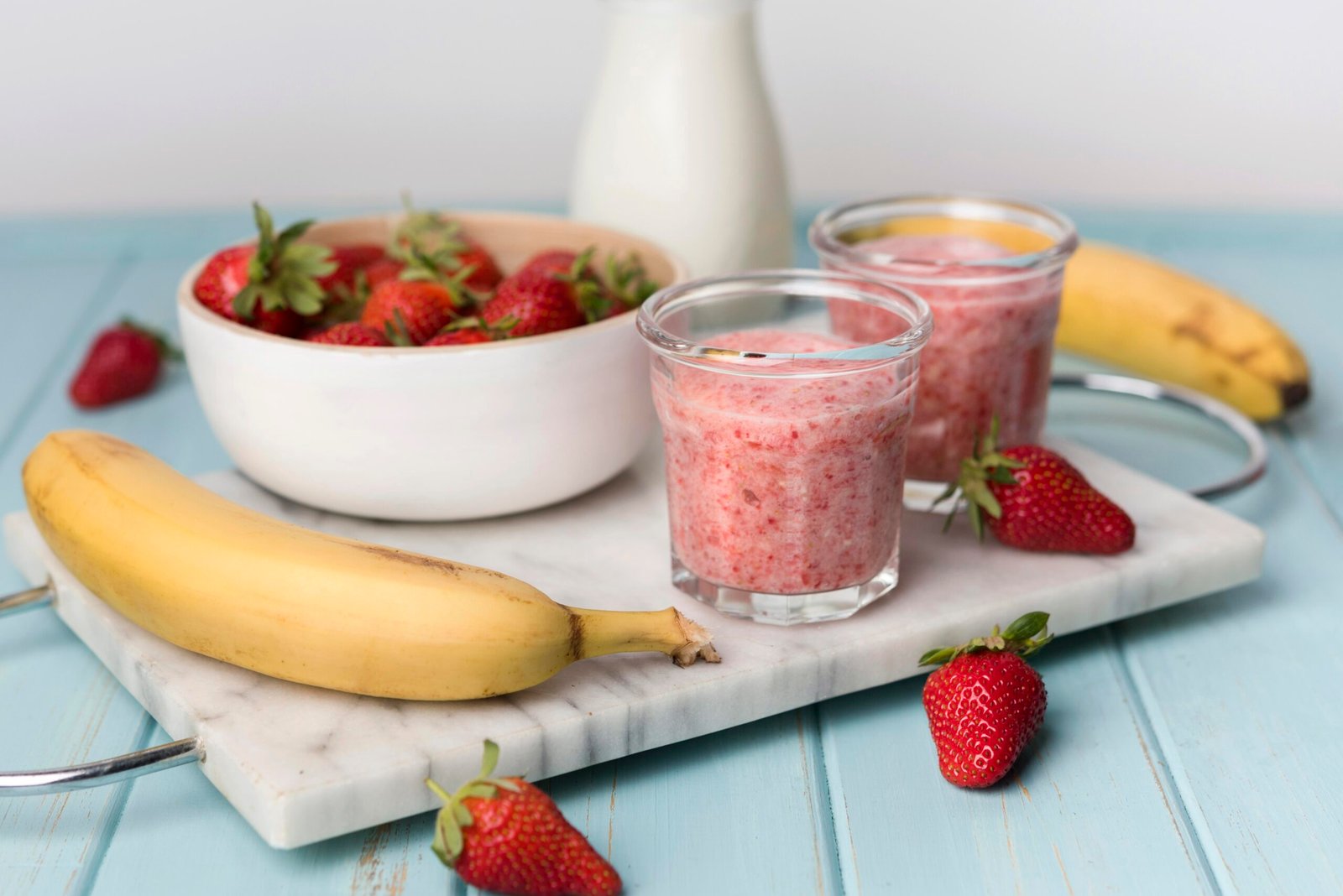 Dos vasos de batido rosa de fresa y plátano sobre una tabla de mármol, rodeados de fresas frescas, un bol con fresas y plátanos.