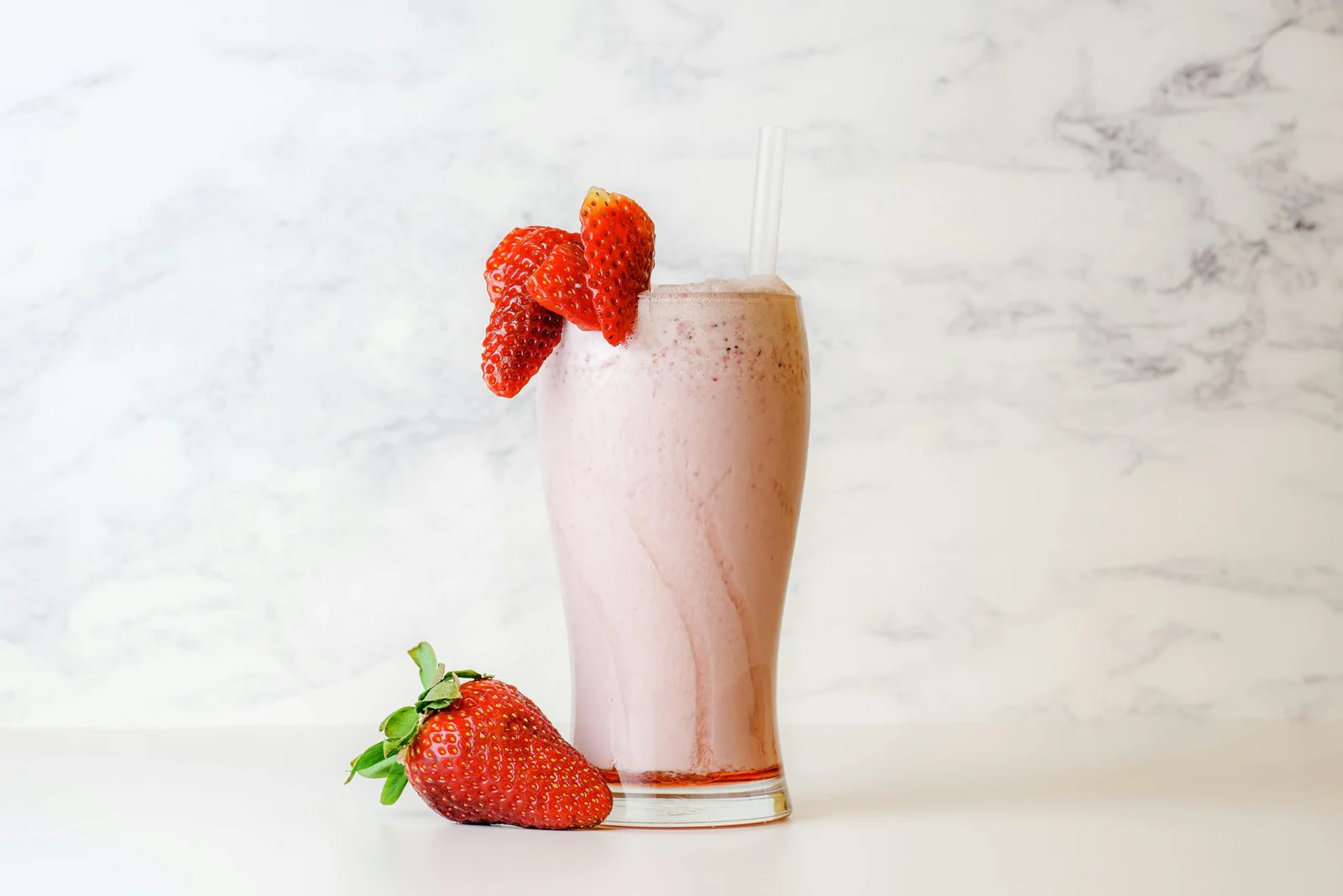 Un vaso alto con un batido cremoso de fresa, decorado con fresas frescas en el borde, situado junto a una fresa entera sobre un fondo de mármol blanco.