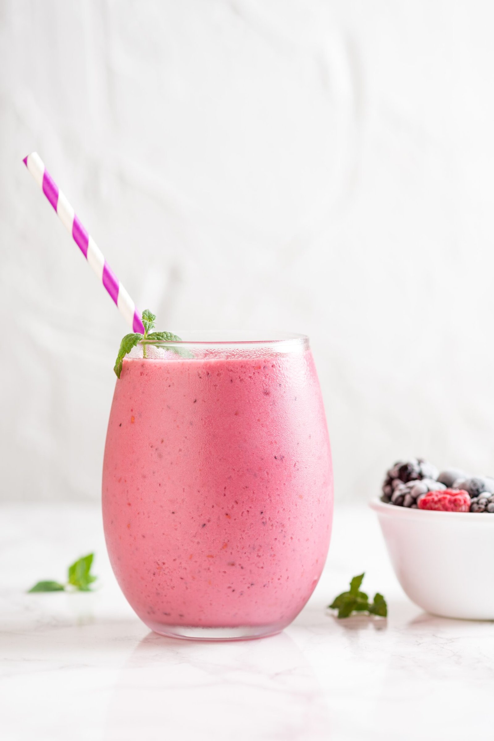 Un vaso de cristal redondeado con un batido saludable rosa vibrante de frutos rojos, decorado con una pajita de rayas blancas y moradas y una hoja de menta, junto a un pequeño cuenco con bayas congeladas.
